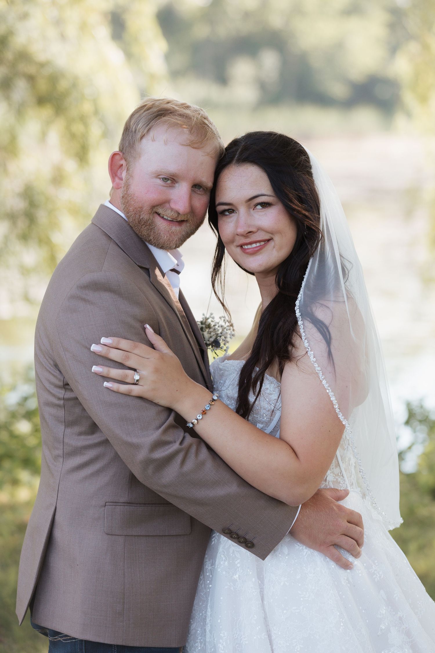 Bride and groom share a romantic kiss on a wooden bridge surrounded by lush green trees on their wedding day.
