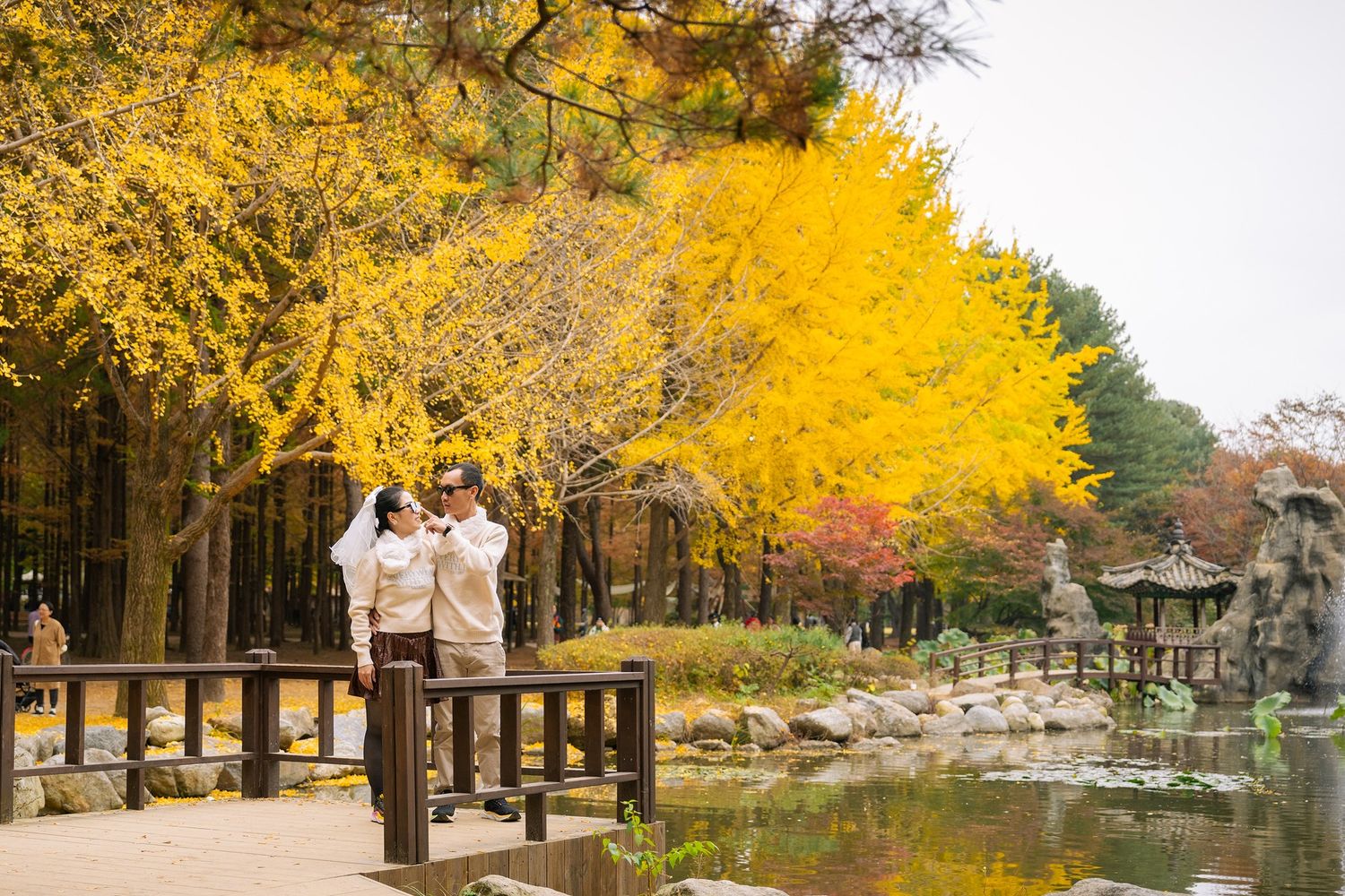 Couple in traditional attire poses by a pond surrounded by vibrant yellow autumn ginkgo trees in a serene park.