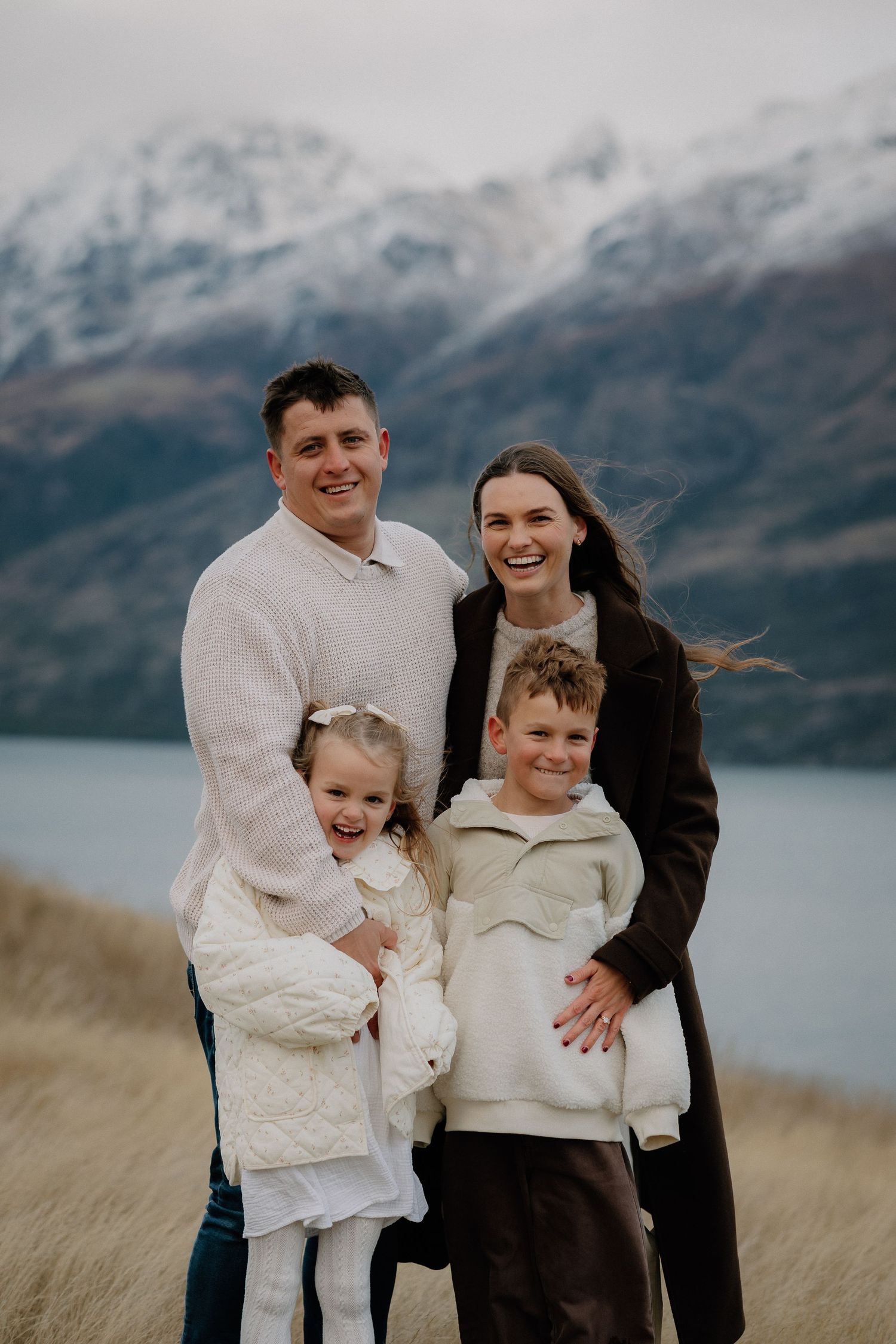 Family of four smiling outdoors with snow-capped mountains and lake in background, New Zealand.