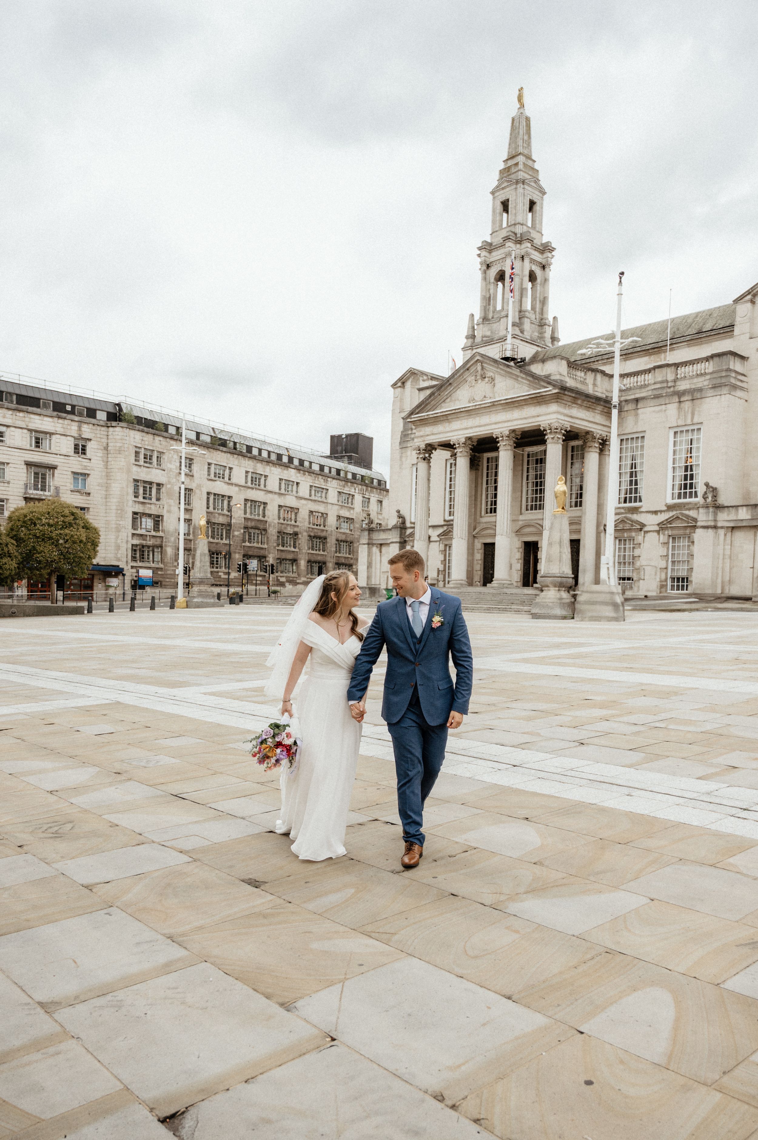 Wedding couple walks across historic stone courtyard with classical architecture.