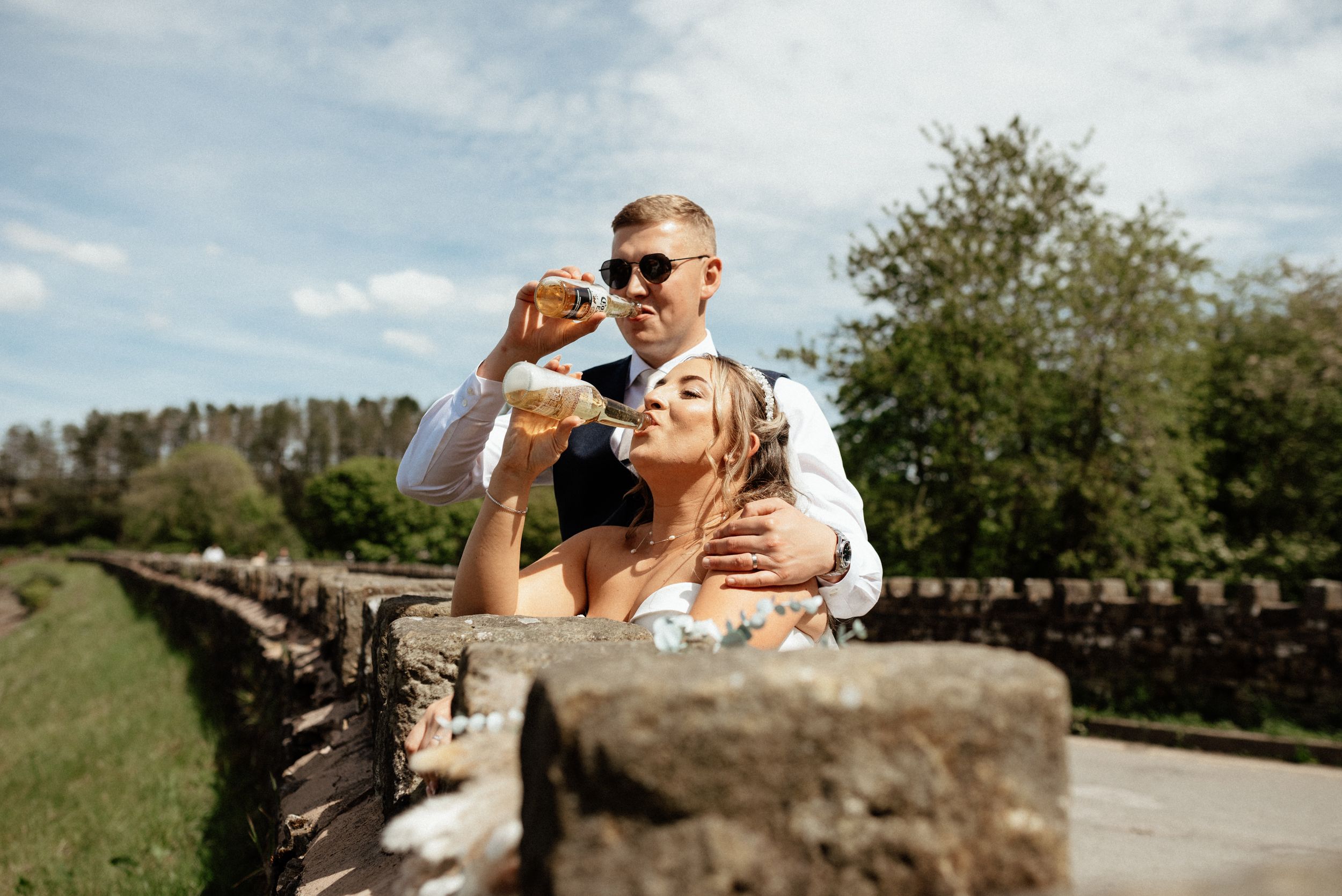 Newlyweds share drinks and laughter on rustic stone wall outdoors.
