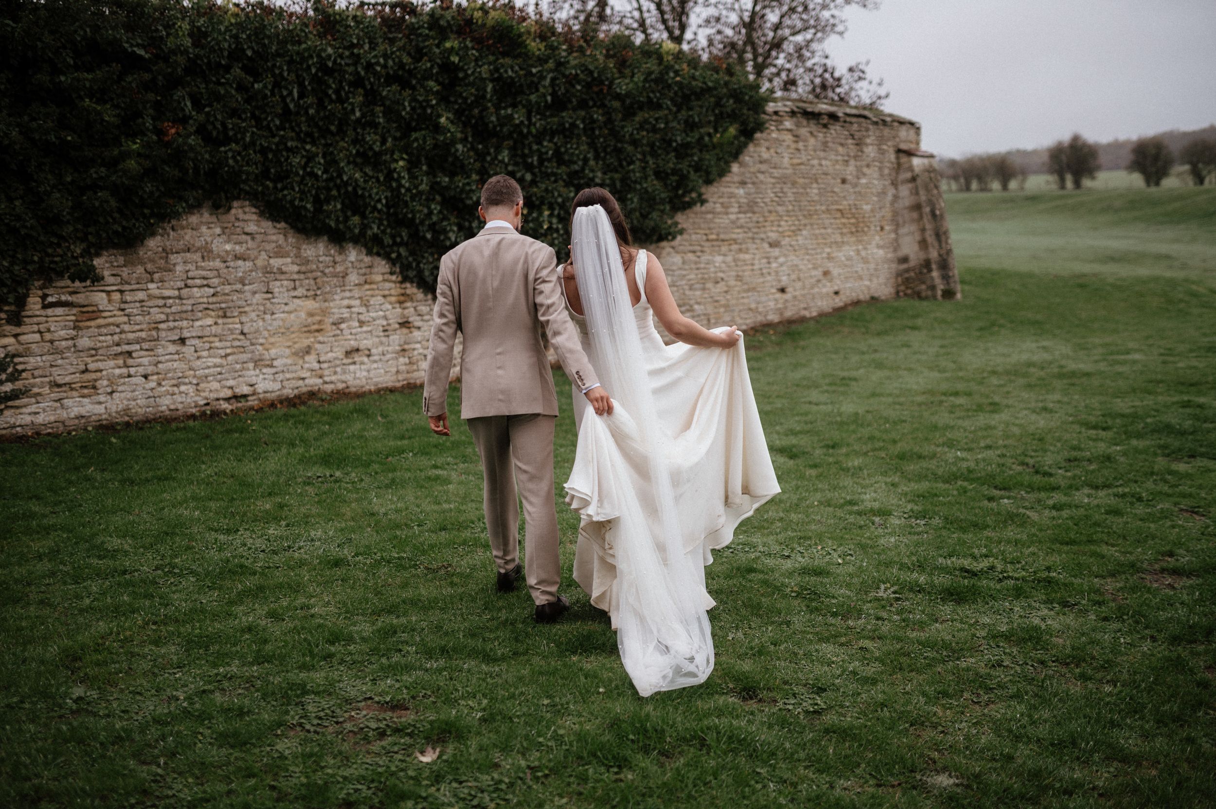 A couple walks along a stone wall on their wedding day, with the bride's white dress flowing behind them on green grass.