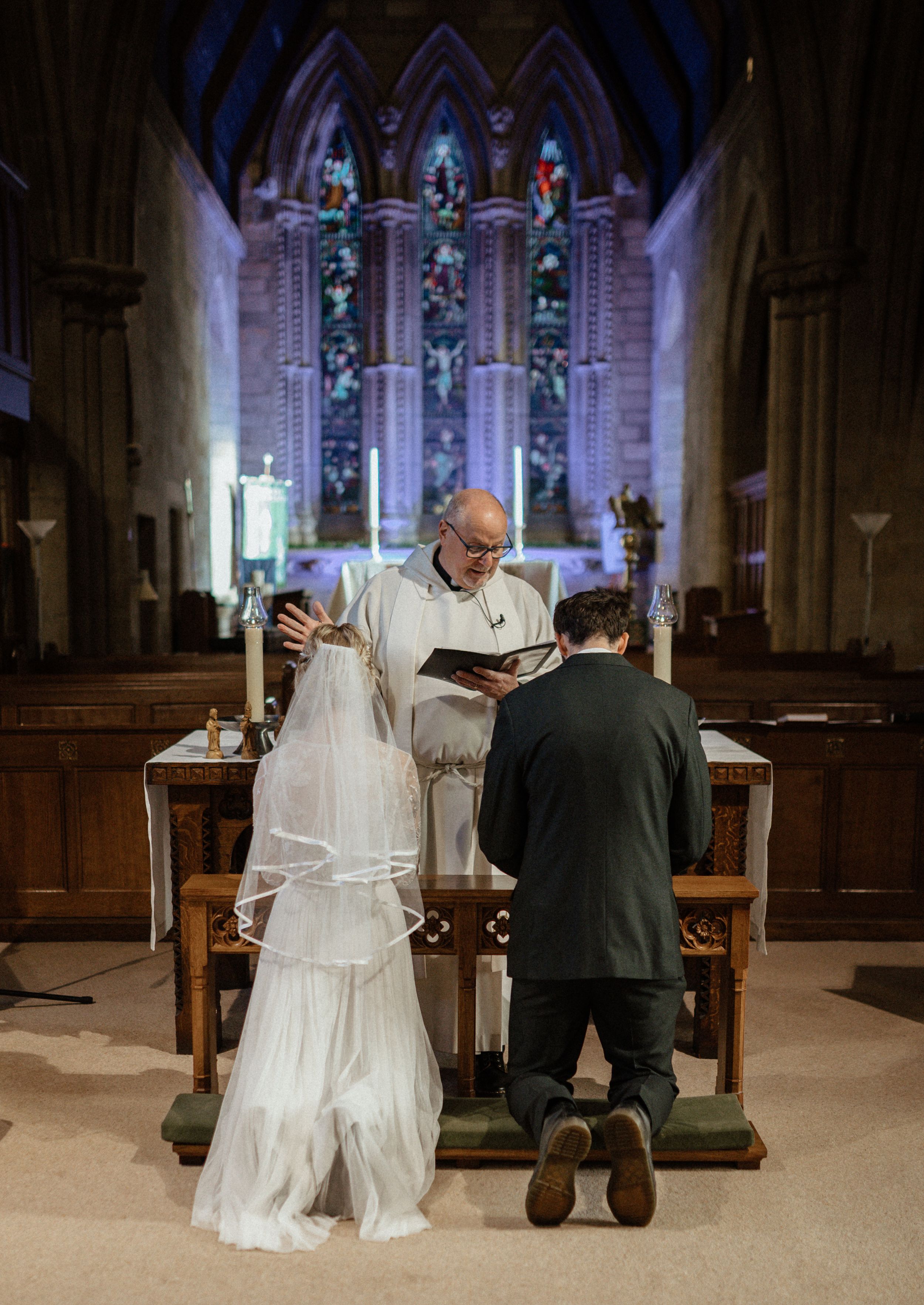 A couple prays together in a church sanctuary with stained glass windows and wooden pews visible in the background.