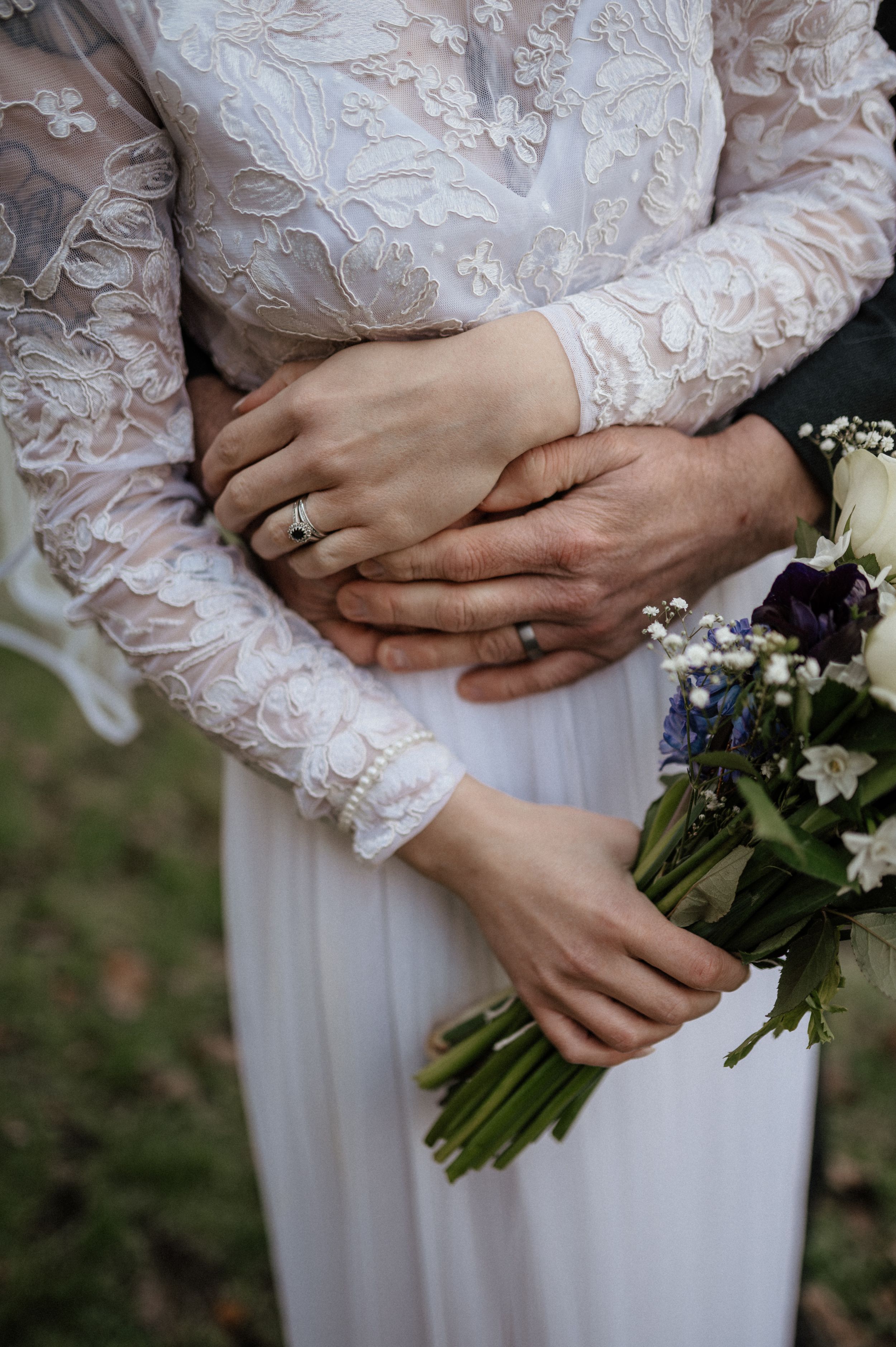 Close-up of hands wearing wedding rings holding a bouquet, with delicate lace sleeves of a white wedding dress.