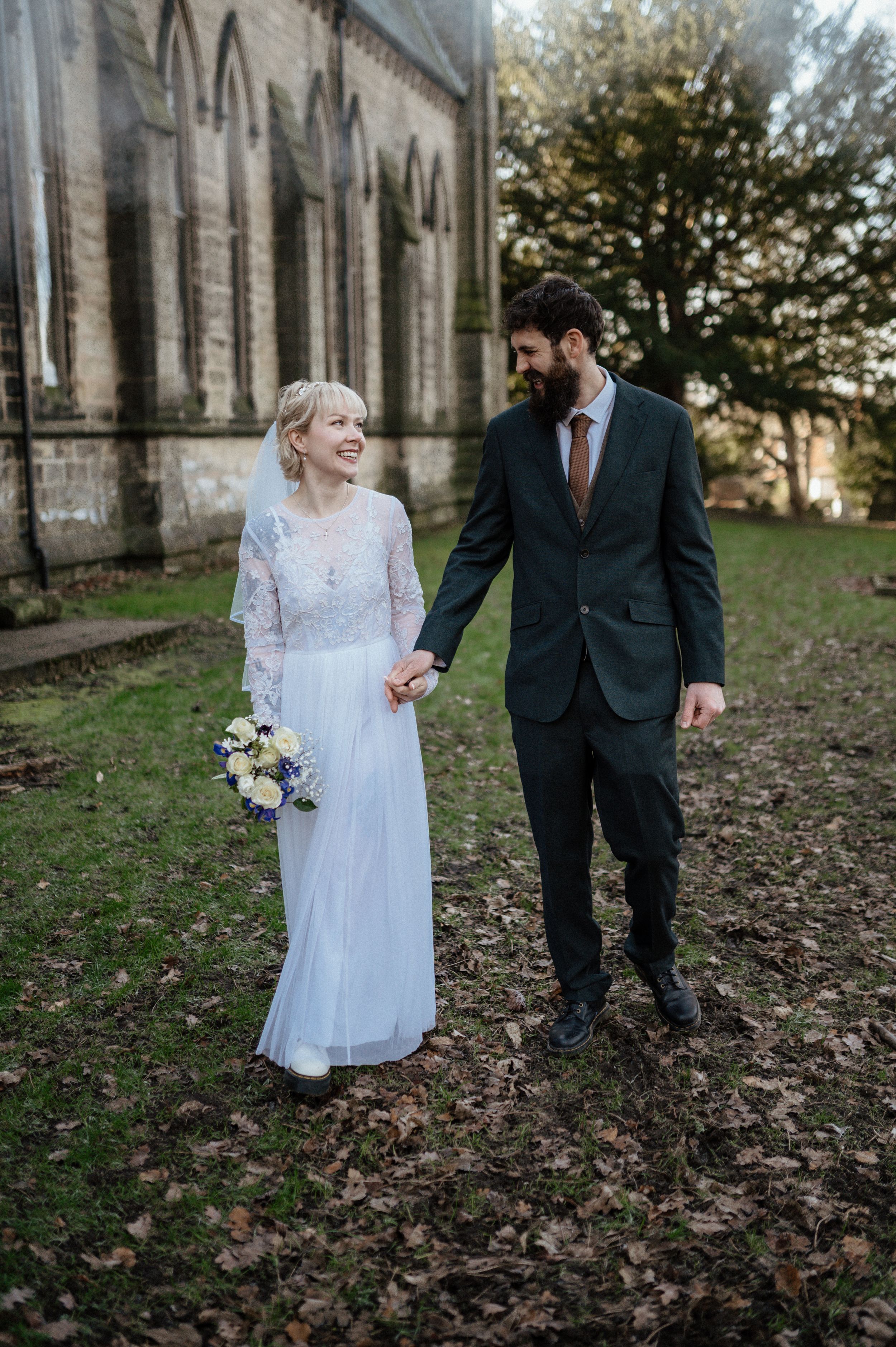 A couple shares a joyful moment walking through autumn leaves outside a historic stone building.