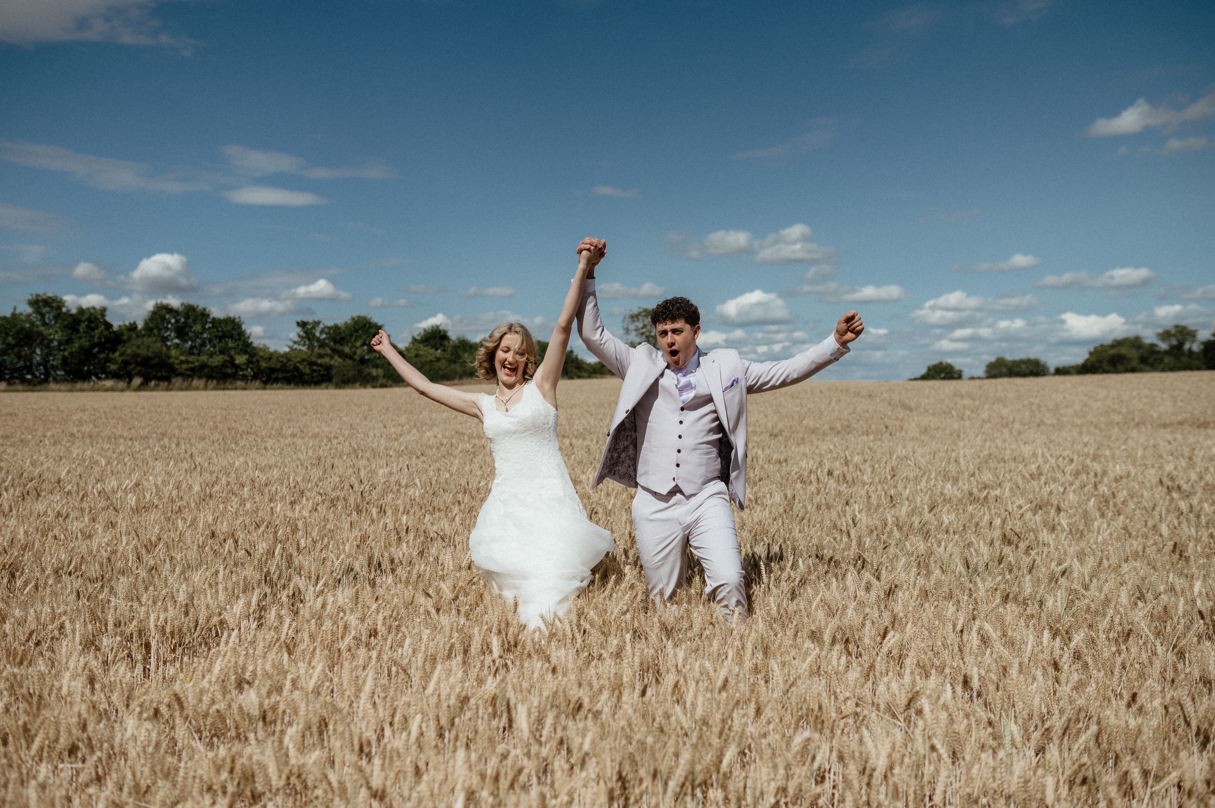 Joyful newlyweds celebrate in a golden wheat field under blue summer sky.