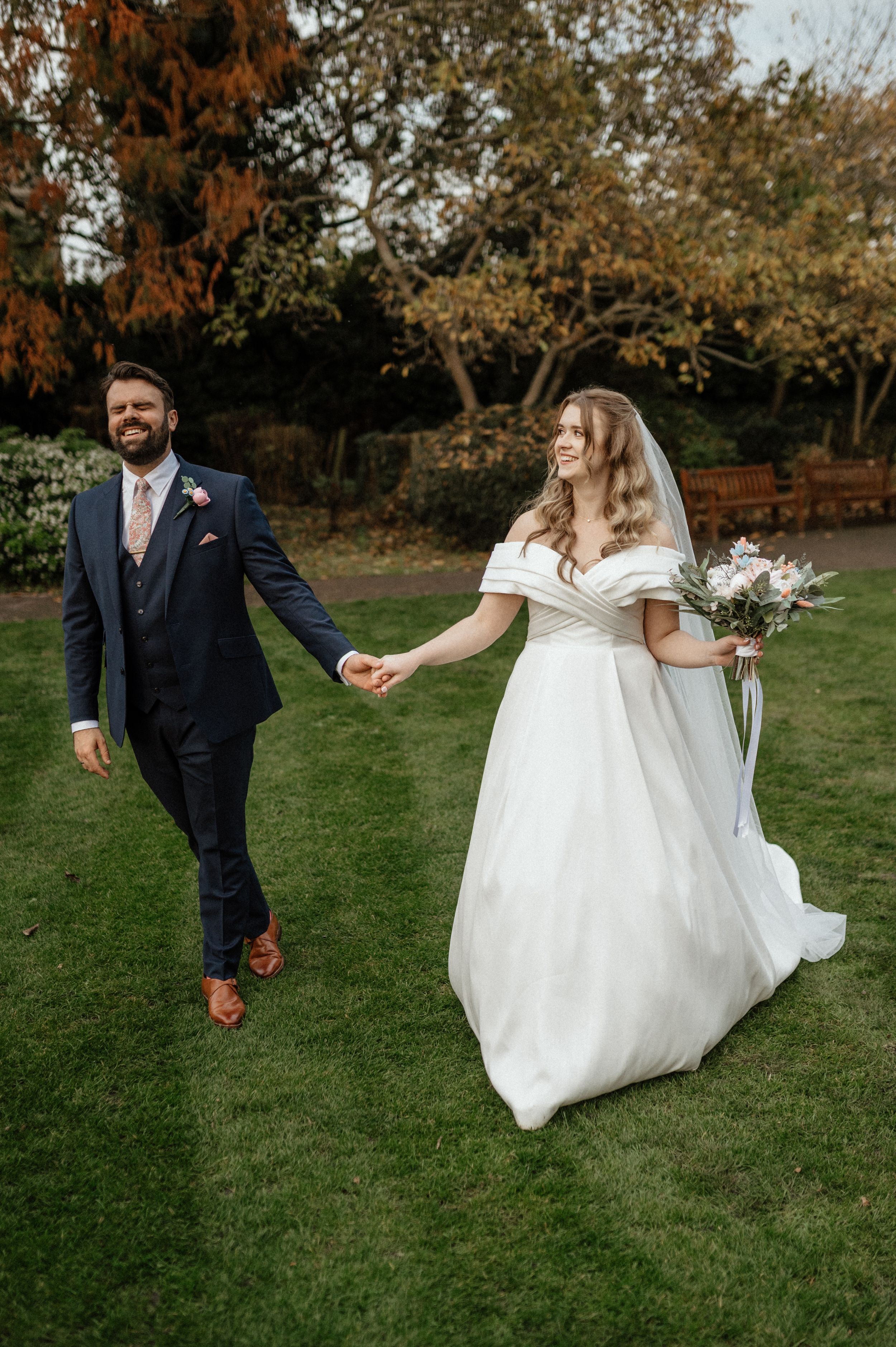 A couple walks hand in hand on a green lawn, with the bride wearing an elegant off-shoulder white gown and holding a bouquet.