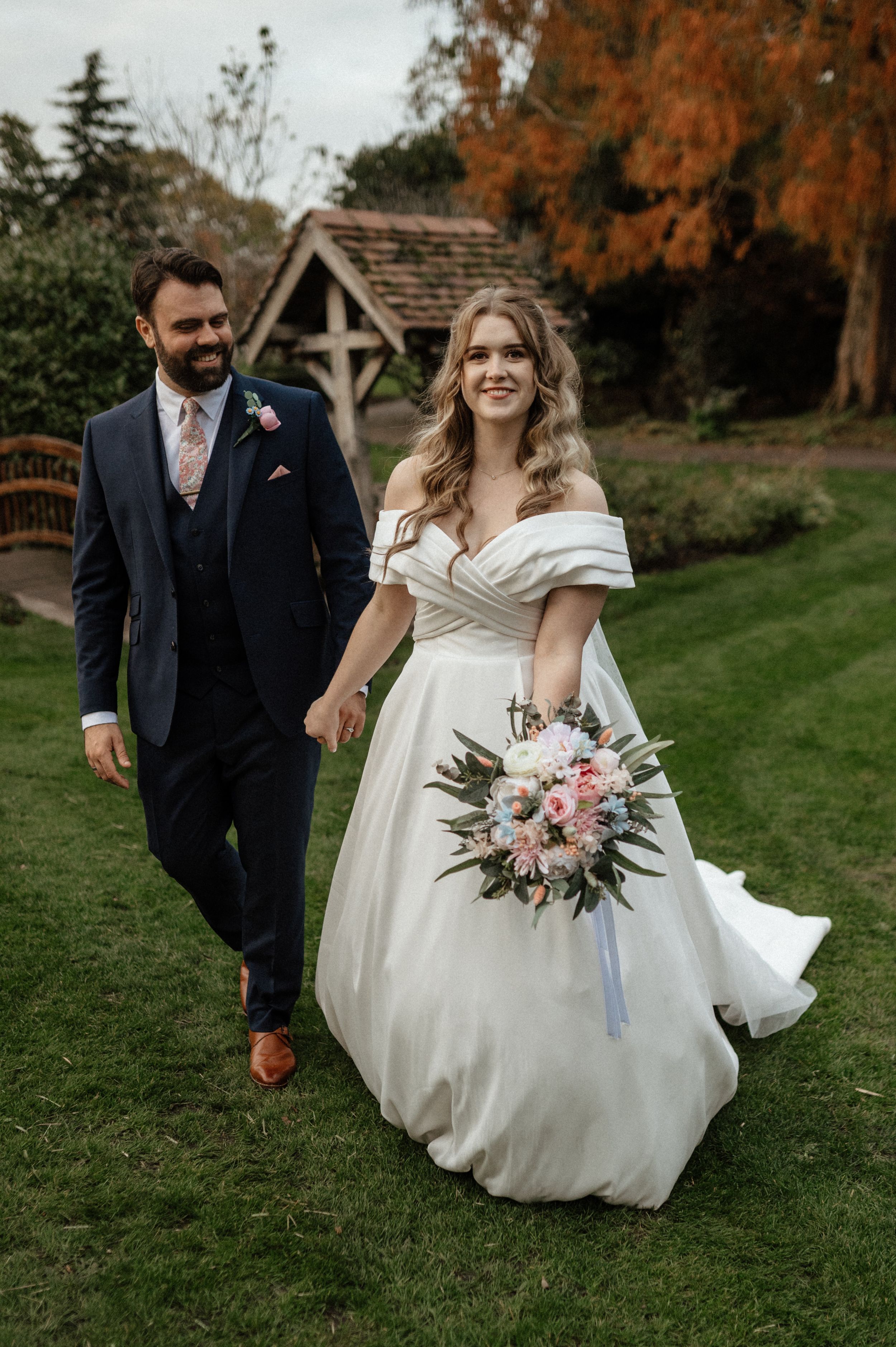 A romantic autumn wedding photoshoot featuring a couple walking together on lush green grass with fall foliage in the background.