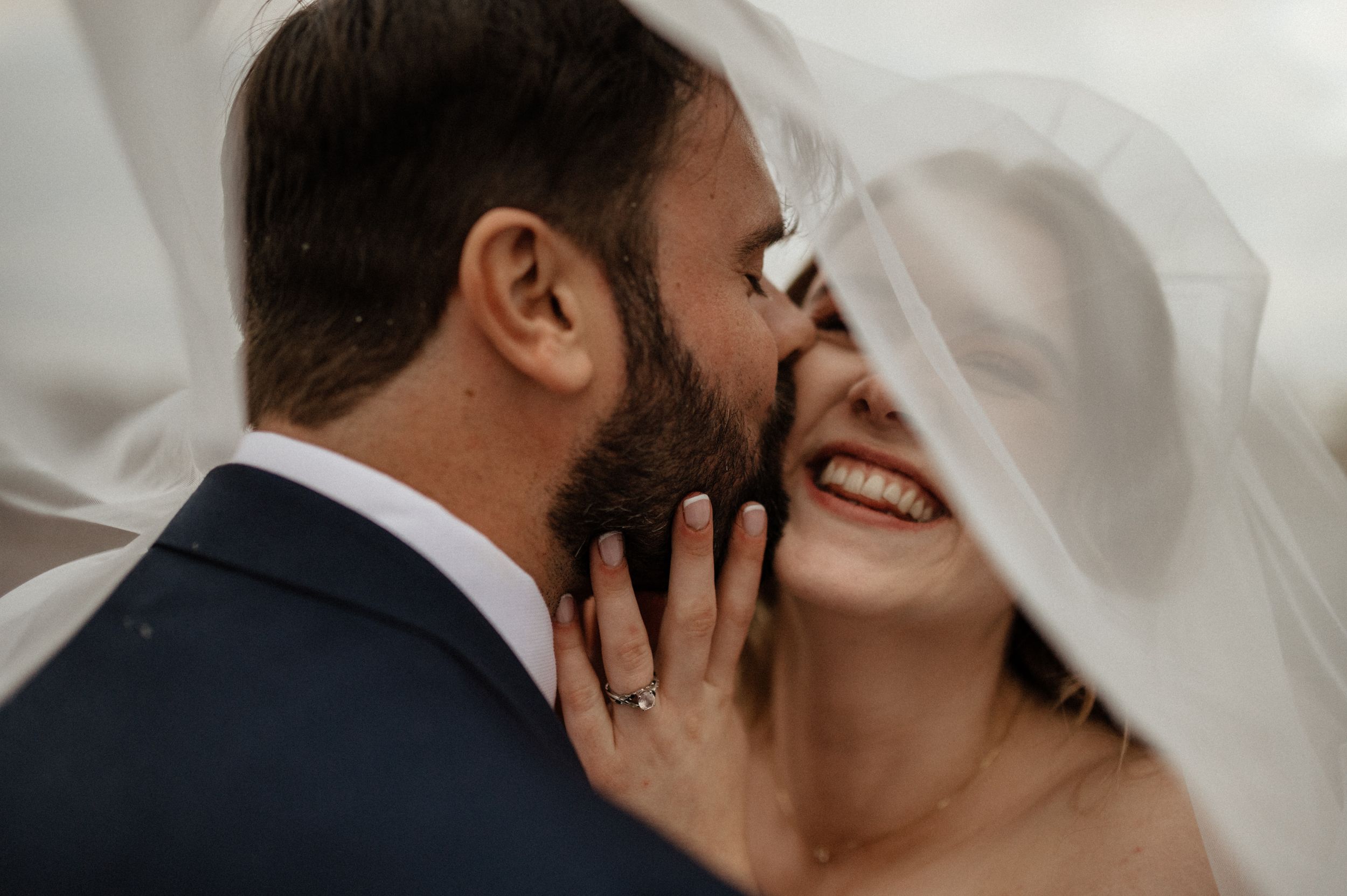 Close up romantic wedding photo of couple kissing under flowing white veil in natural light.