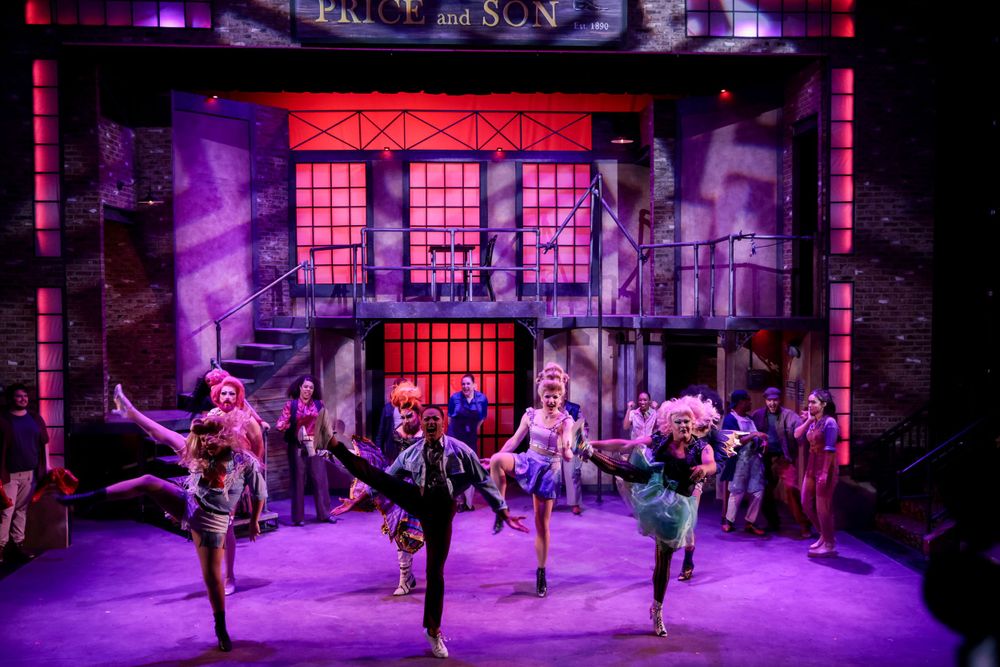 Theatrical performers dancing on stage with dramatic purple lighting and urban scenery backdrop.
