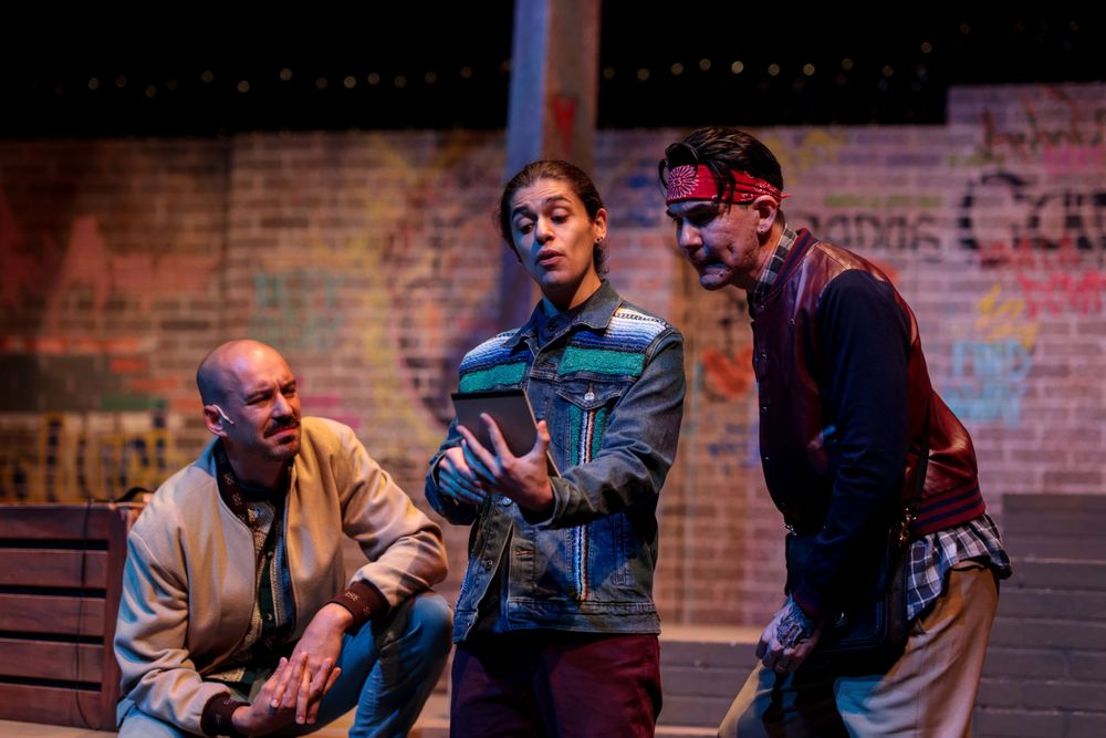 Three performers rehearsing or acting in a dimly lit theater space with brick wall backdrop.