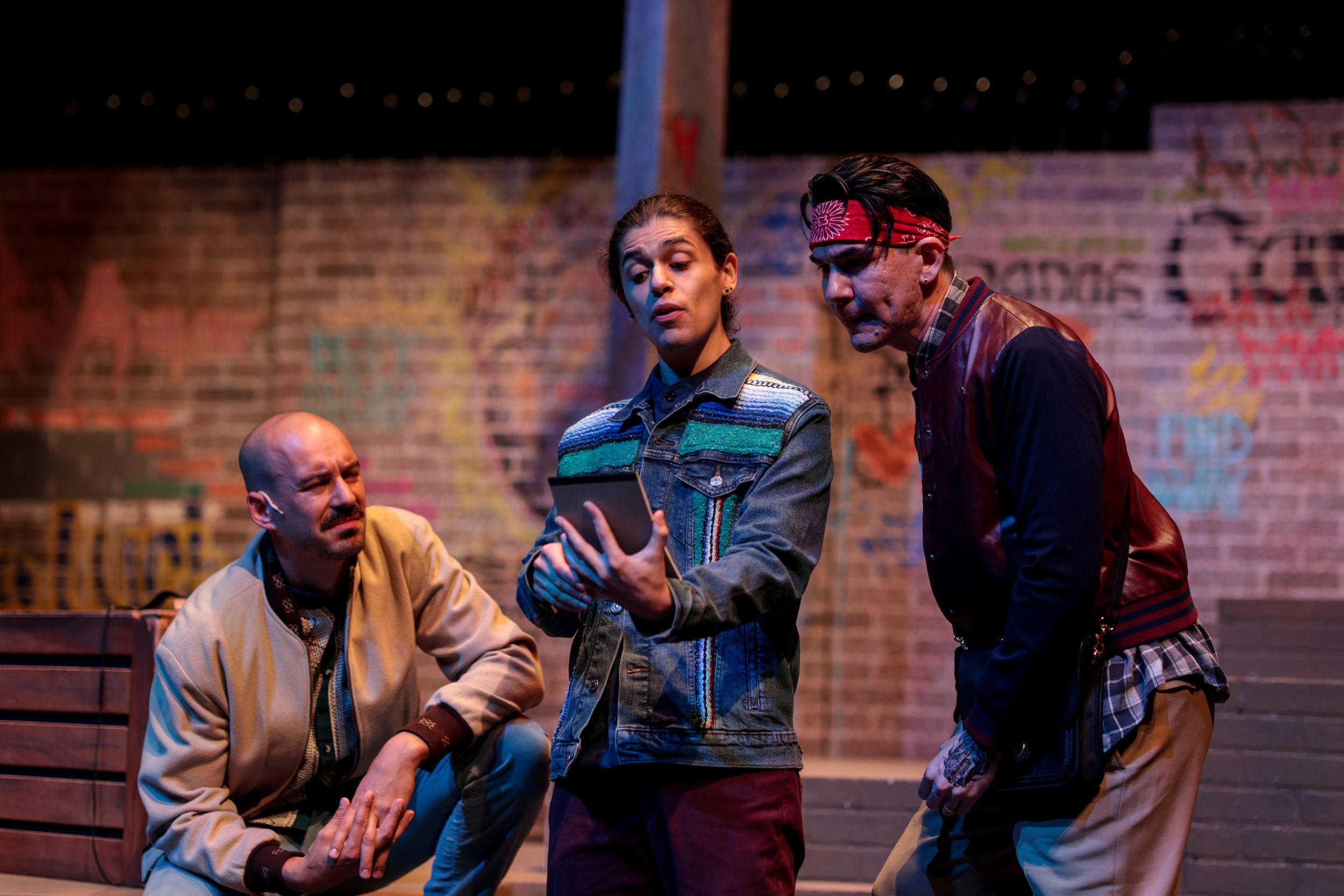Three performers rehearsing or acting in a dimly lit theater space with brick wall backdrop.