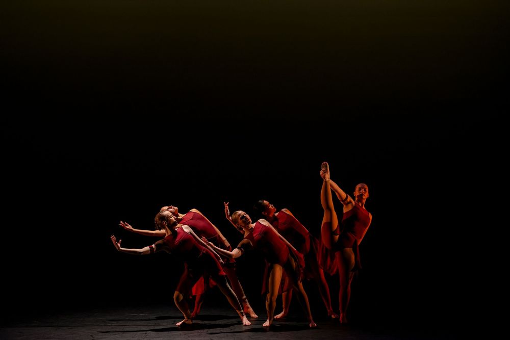 A dramatic modern dance performance sequence showing dancers in red costumes moving across a dark stage. Theatre photography.