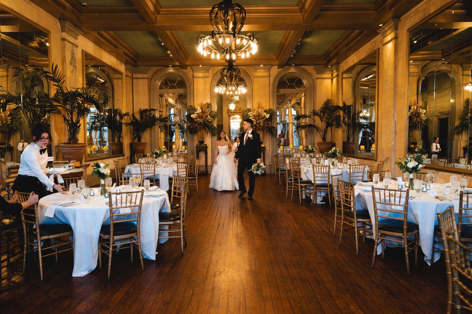 Bride and groom stand in elegant wedding reception hall with chandelier, white tablecloths, and gold chiavari chairs.