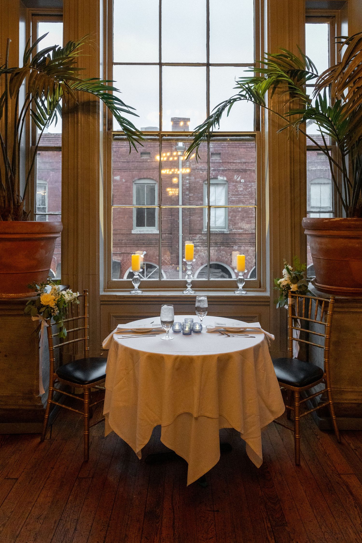 Elegant restaurant table for two with white linen, candles, and orange drinks by a large window overlooking a brick building.