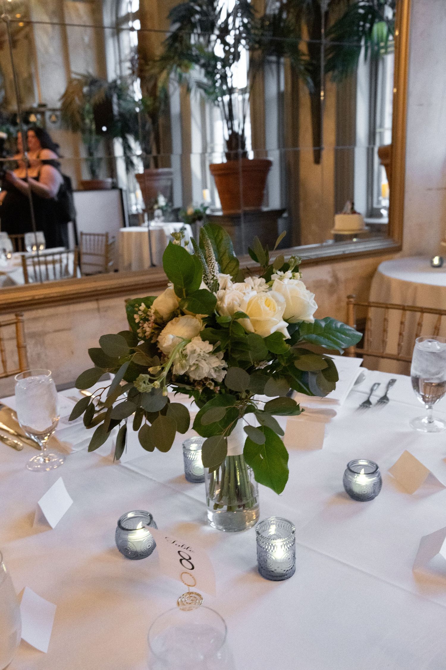 Elegant wedding table centerpiece with white roses, greenery, and candles on a white linen tablecloth.