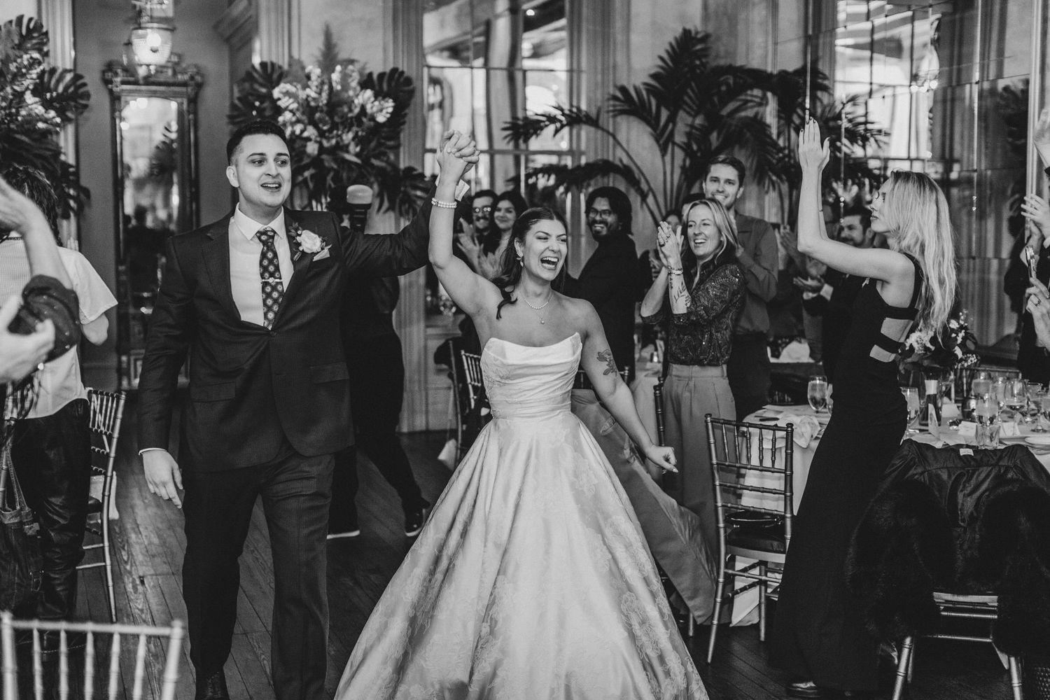 Black and white photo of a joyful bride and groom entering their wedding reception as guests celebrate around them.