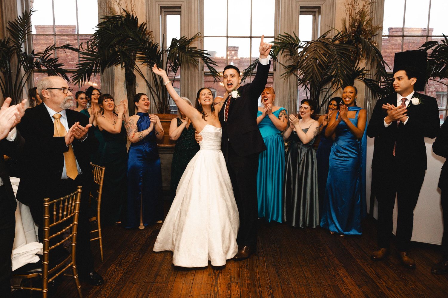 Bride and groom celebrate grand entrance at wedding reception with guests in blue gowns clapping in elegant venue.