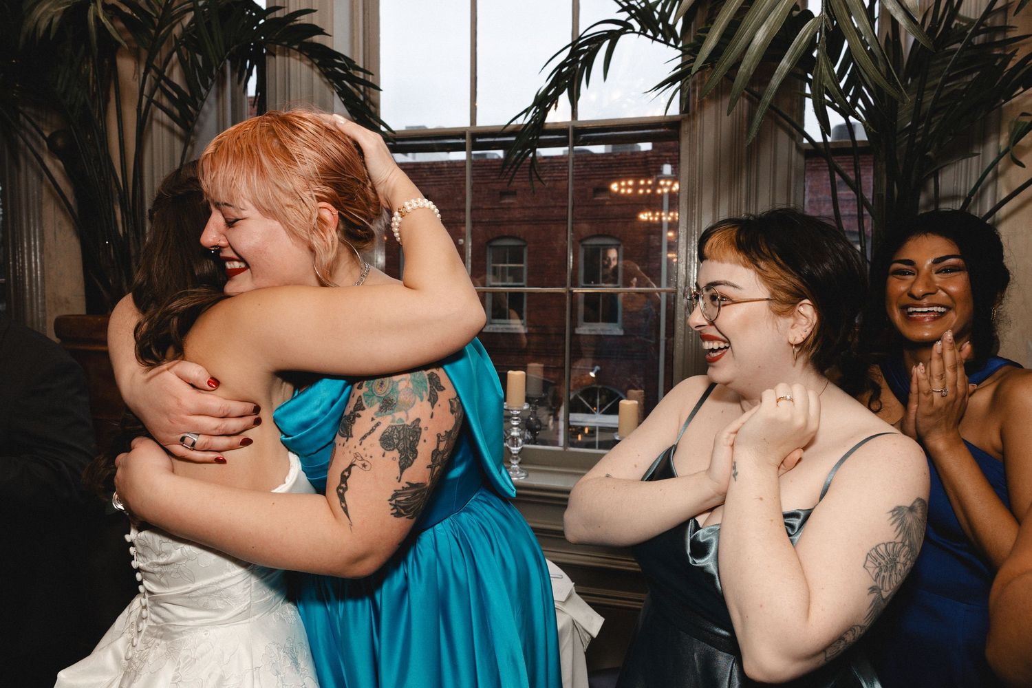 Bride hugging bridesmaid in teal dress at wedding reception while guests laugh and celebrate nearby.