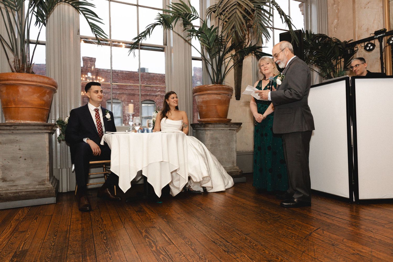 Bride and groom seated at sweetheart table during indoor wedding ceremony with officiant reading vows.