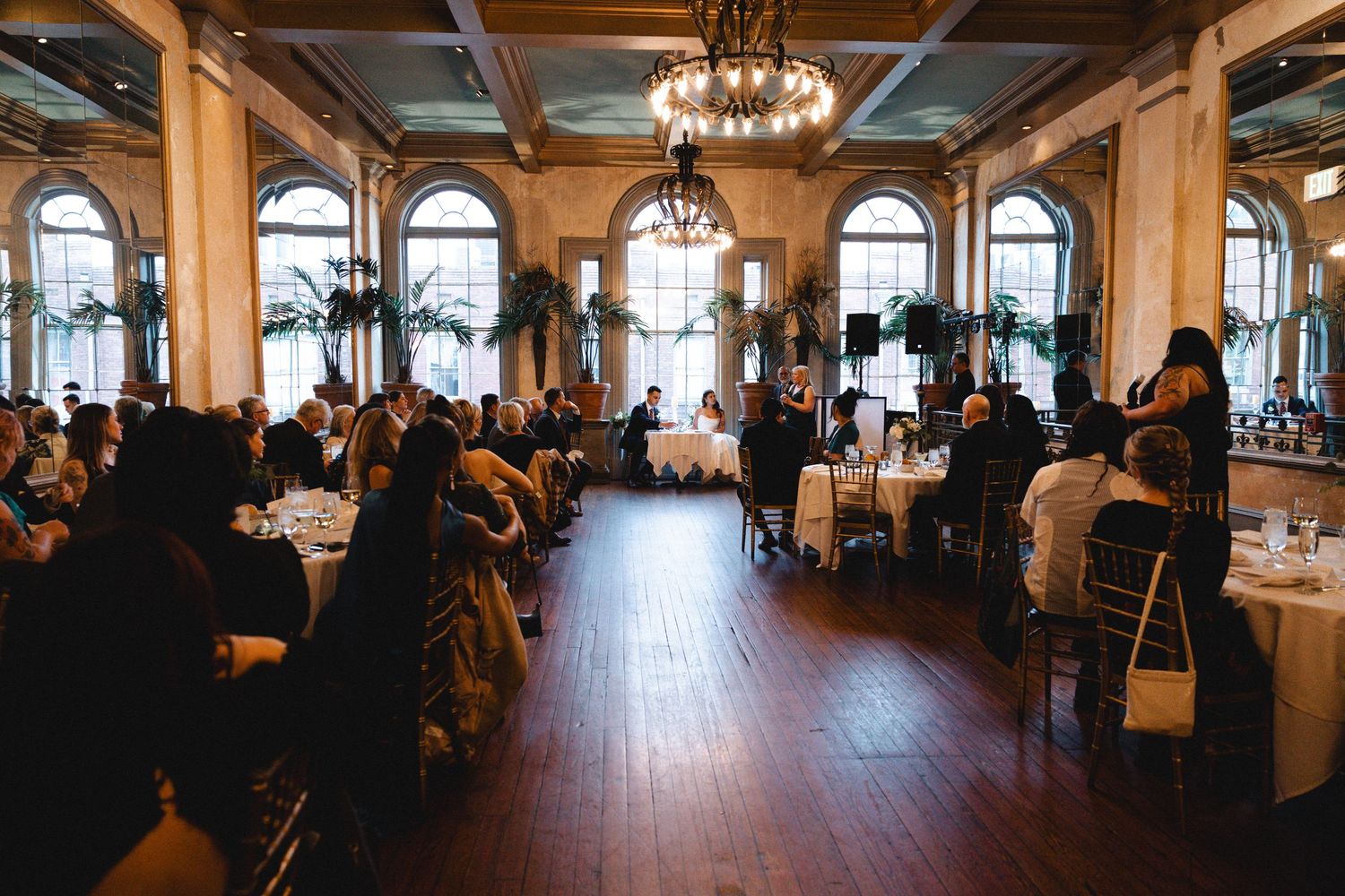 Grand wedding reception hall with arched windows, chandelier, and guests seated at round tables.