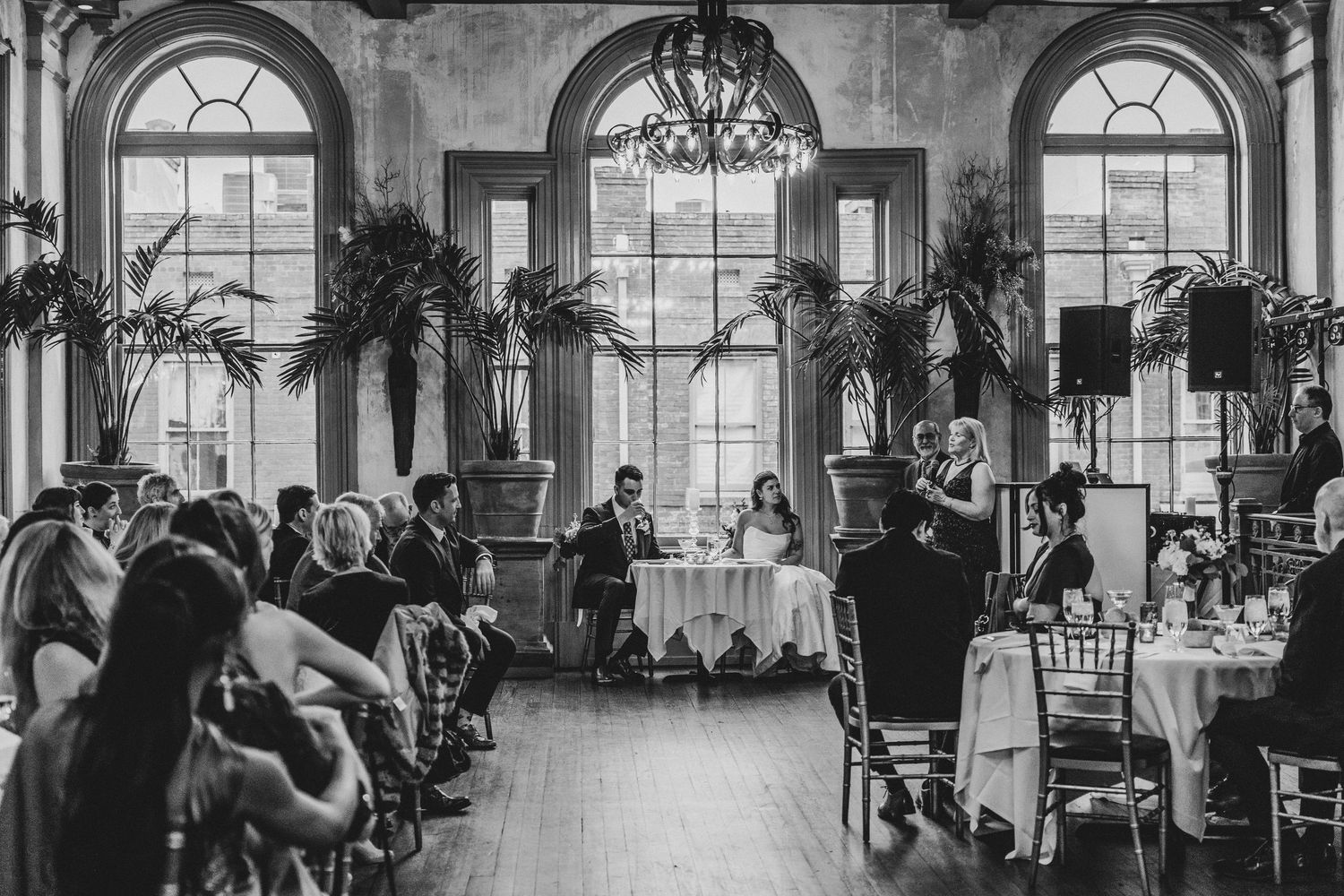 Black and white photo of an elegant wedding reception in a grand ballroom with tall arched windows and chandeliers.