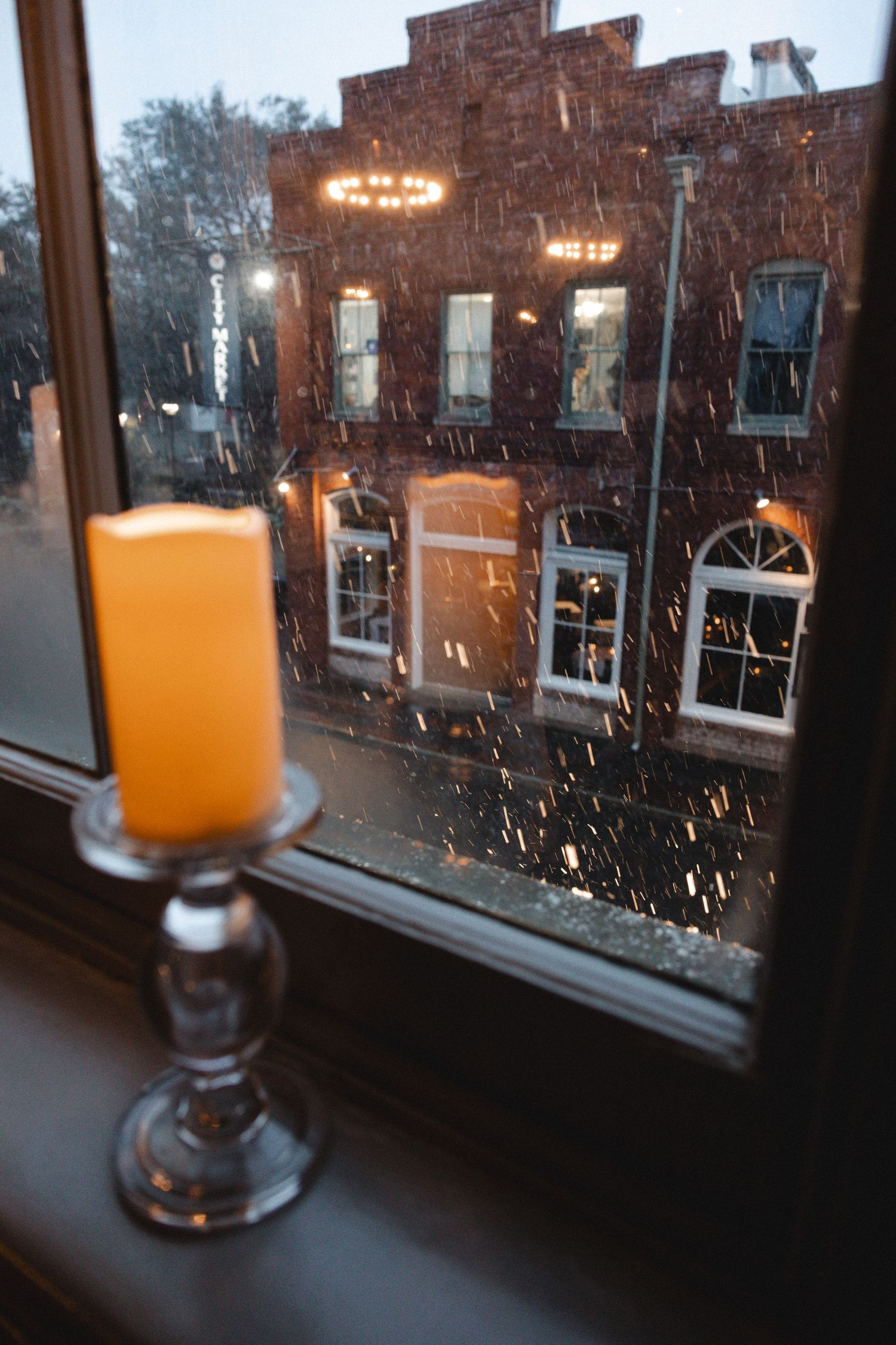 A glowing candle on a silver holder sits on a windowsill overlooking snowy brick buildings at dusk.