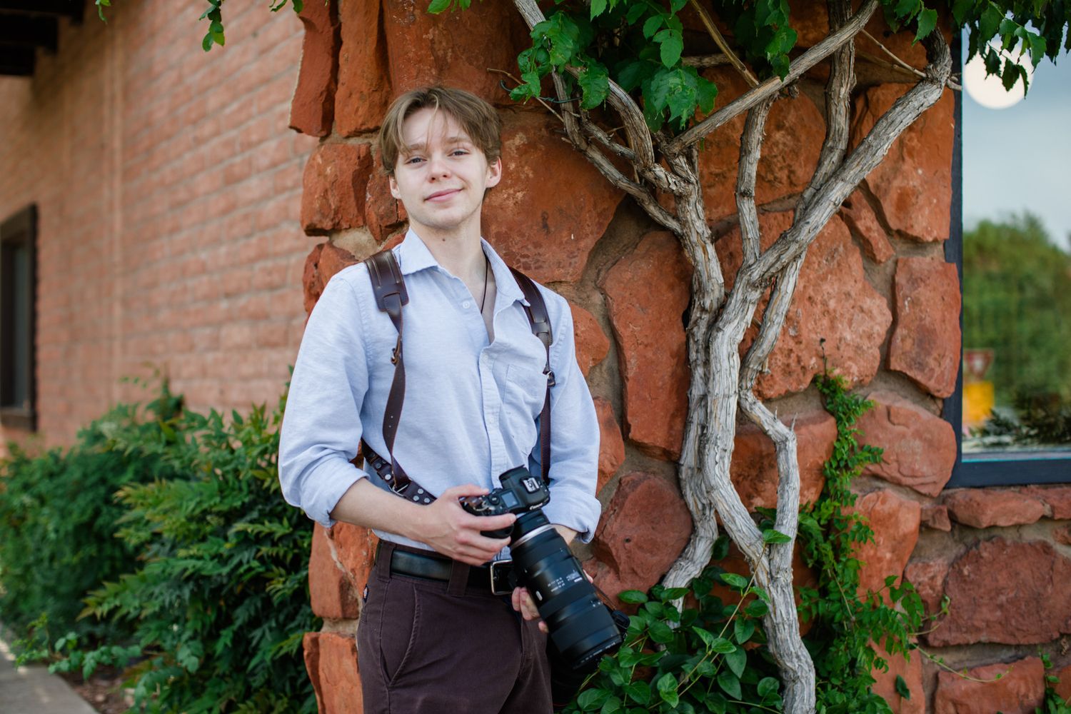 Chandler wearing a camera harness outdoors near a red stone wall with green foliage. He holds a canon R5 with a telephoto lens in one hand.