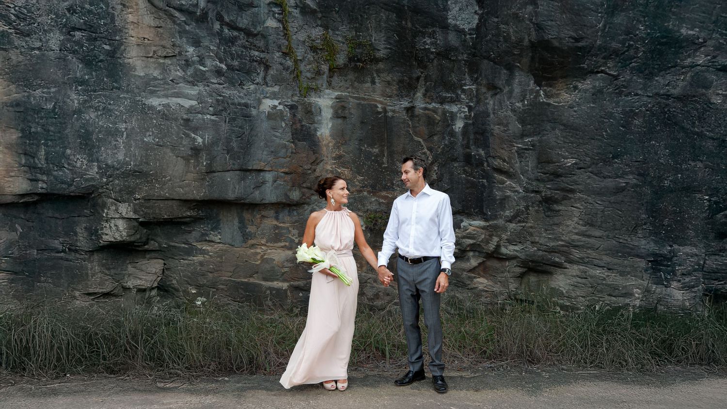 Bride holding calla lilies and groom holding hands in front of a dramatic rock face, by a Suffolk wedding photographer.