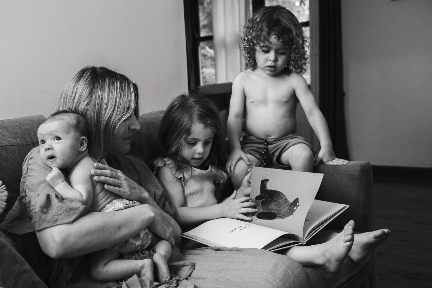 Candid black and white lifestyle family photography in Suffolk of a mother with her newborn baby, young daughter, and toddler son reading.