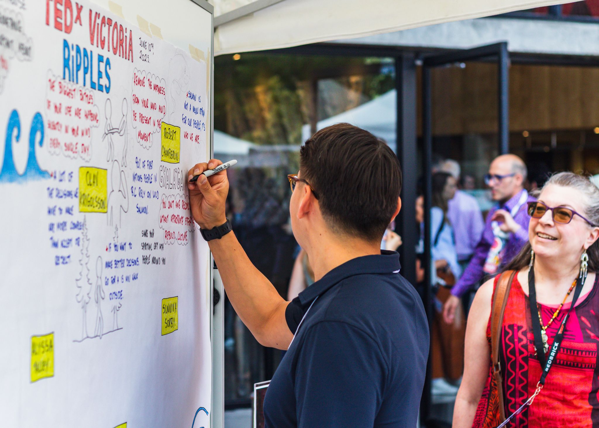 Person writing on a TEDx Victoria Ripples whiteboard display at an outdoor event with attendees nearby.