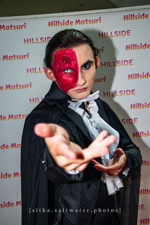 Person in Phantom of the Opera cosplay with red mask makeup reaching toward camera at Hillside Matsuri event.