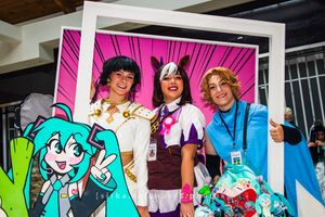 Three cosplayers pose in a decorative anime-themed photo frame at a comic convention.