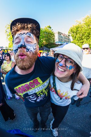 Two face-painted festival-goers smile and pose together outdoors at a lively street fair on a sunny day.