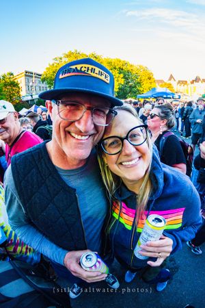 A smiling couple poses together at an outdoor festival, holding canned drinks, wearing glasses and casual clothing.