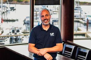 Man in navy polo shirt smiling at marina restaurant with boats visible through large windows behind him.
