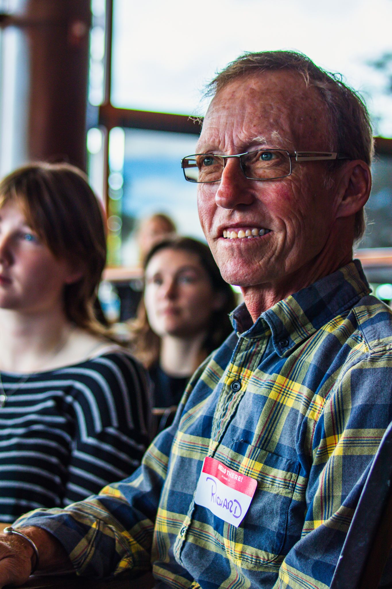 Smiling middle-aged man wearing glasses and plaid shirt with name tag sits among audience members at an indoor event.