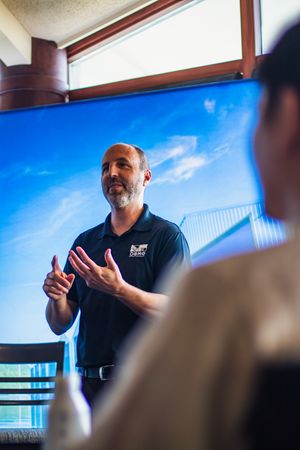 A bearded man in a black polo shirt presents to an audience in front of a large blue sky display screen.