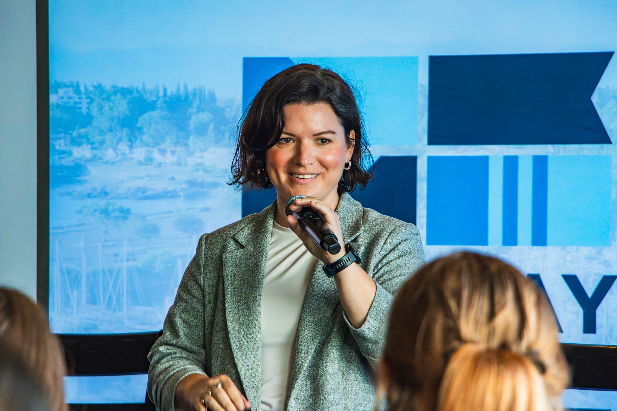 A smiling woman speaker holds a microphone while presenting in front of a blue branded backdrop.