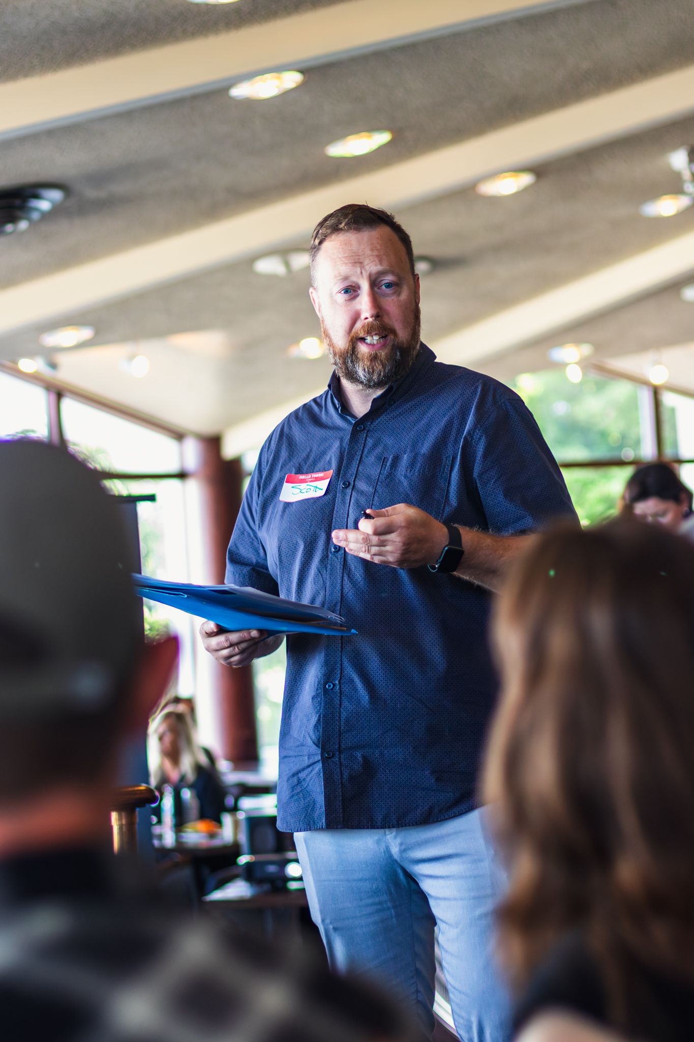 Bearded man in blue shirt holds folder while speaking to audience in casual restaurant meeting setting.