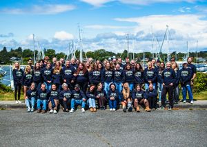 Large group of people in matching navy hoodies posing outdoors near a marina with sailboats and blue sky.