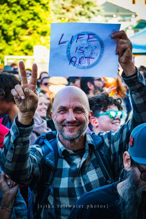 Smiling bald man holds a Life is Art sign and flashes a peace sign at an outdoor festival crowd.