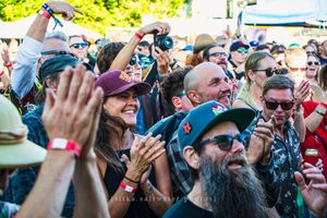 Crowd of happy festival-goers clapping and cheering outdoors, wearing hats and sunglasses on a sunny day.