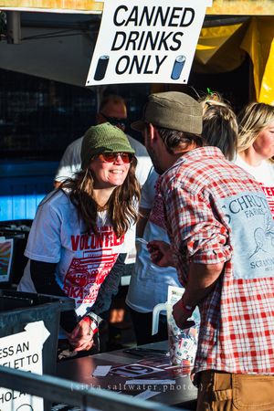 Smiling female volunteer at outdoor event booth with Canned Drinks Only sign, wearing green beanie and red volunteer shirt.
