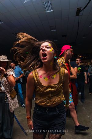Woman with neon face paint dancing energetically at a festival event, hair flying, wearing a gold top.