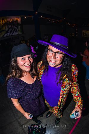 Two festively dressed people pose together at a party, one in a purple hat and colorful jacket, the other in a dark outfit.