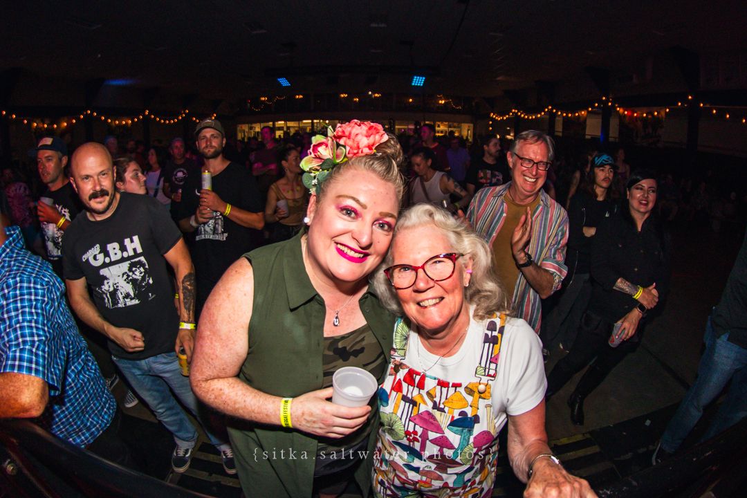 Two women smiling and posing together at a lively indoor event with colorful lights and crowd.