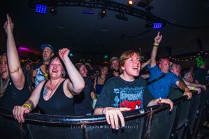 Excited concert crowd cheering and raising hands at an indoor music venue with colorful stage lighting.