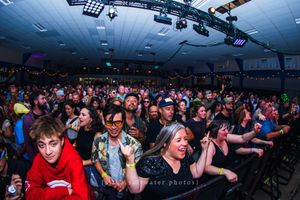 A large crowd of excited concert-goers enjoying a live music event inside a dimly lit venue with stage lighting.