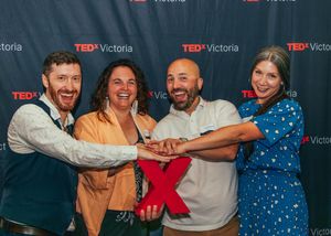 Four smiling people pose together at a TEDxVictoria event, joining hands in front of a branded backdrop.